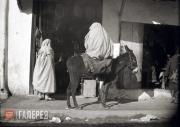 Two figures in burnouses, with a donkey in front of a shop cafe Two figures in burnouses, with a donkey in front of a shop cafe